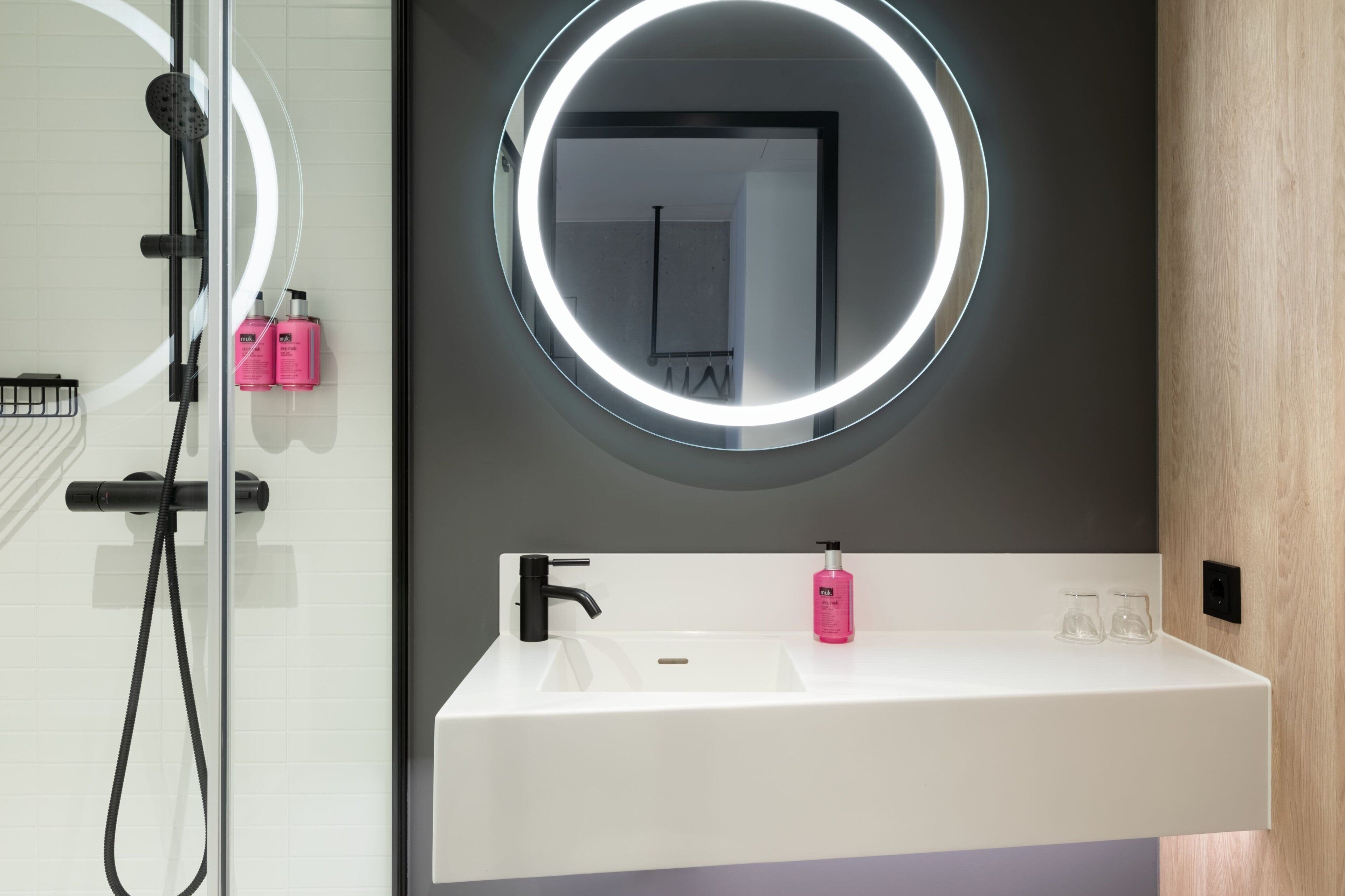 A modern bathroom with a white sink, a round mirror with a light halo, and a shower area.