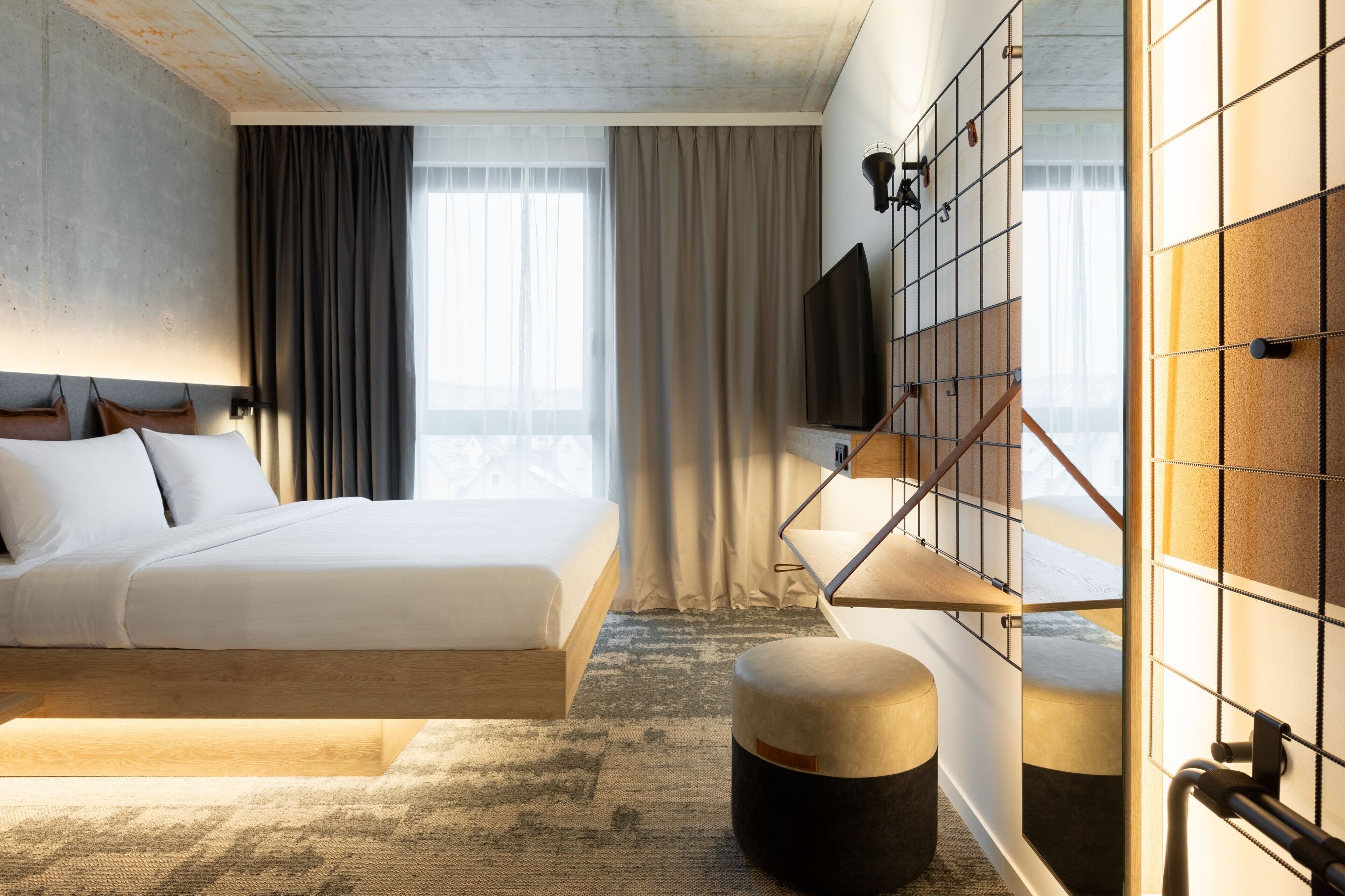 A modern hotel room with a large bed, a flat-screen TV mounted on a grid wall, and a view of the outdoors.