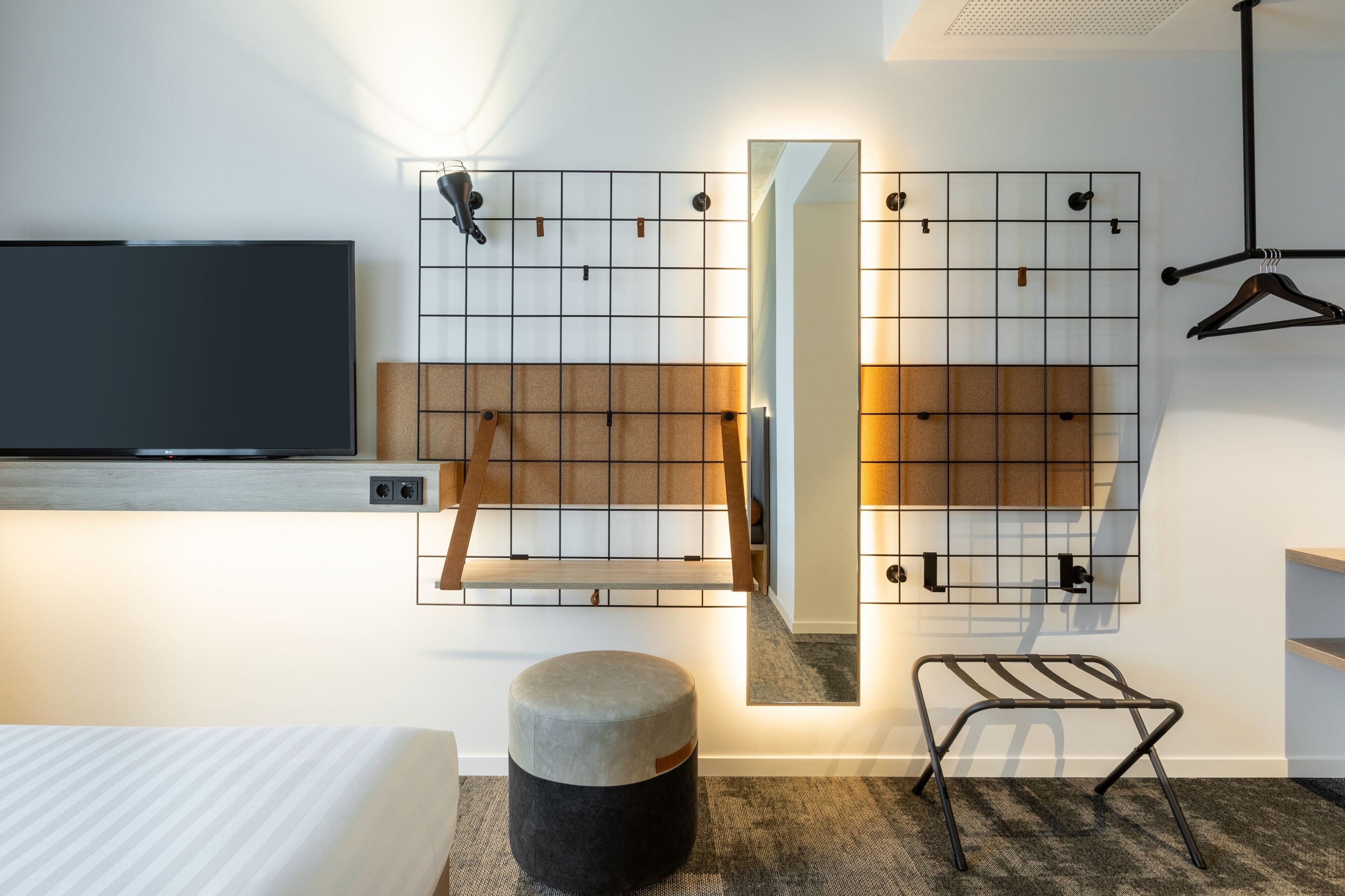A modern hotel room with a flat-screen TV, a large mirror, a wooden shelf, a hanging rack, and a round ottoman.