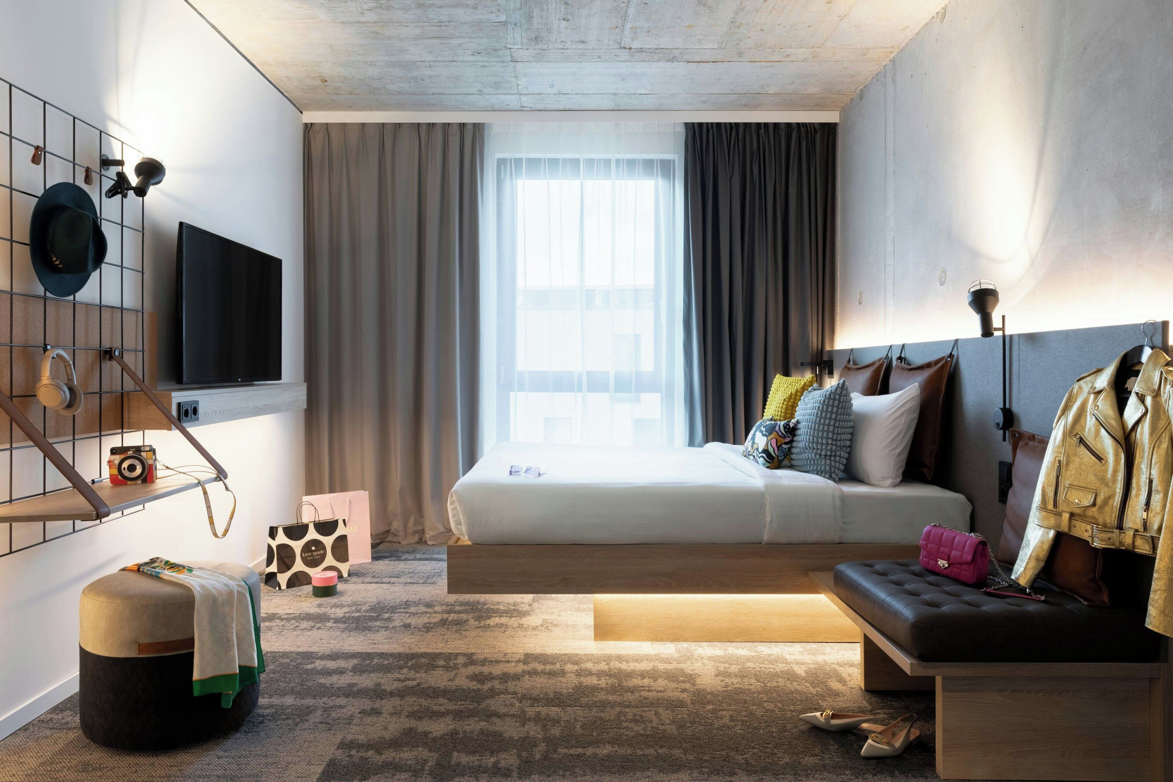 A modern hotel room with a bed, a seating area, a television, and a view of the city.