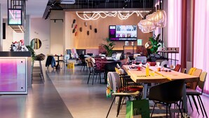 A modern hotel lobby with a long reception desk, seating area with tables and chairs, and a large TV screen displaying content. - Moxy Outletcity Metzingen (Metzingen)