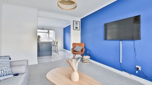 A modern living room with a blue accent wall, a flat-screen TV mounted on the wall, a wooden coffee table with a vase, and a grey sofa. - Spacious 3 Bedroom House in Barry (Barry)