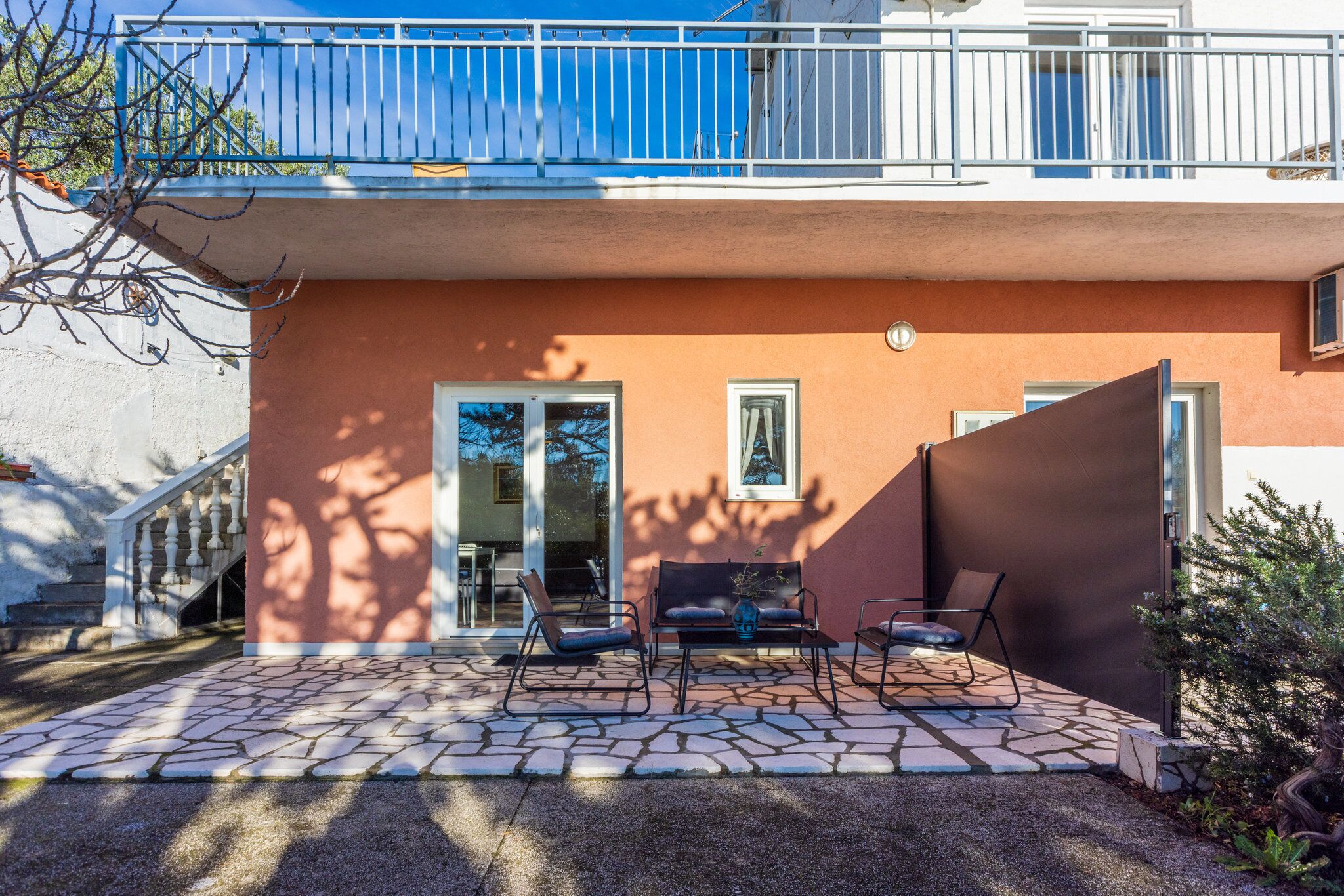 Ein modernes Gebäude mit einer gepflasterten Terrasse, Sitzgelegenheiten im Freien und einem blauen Balkon.