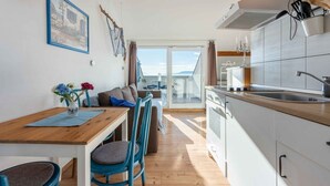 Eine moderne Küche mit weißen Schränken, einer Holz Arbeitsplatte und einem Spülbecken. Ein Essbereich mit einem Holztisch und blauen Stühlen lädt zum Verweilen ein. Ein großes Fenster bietet einen Blick ins Freie.