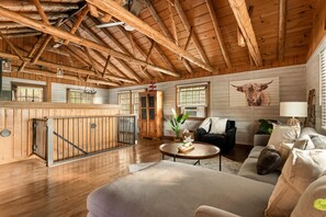 Ein geräumiger Raum mit hoher Decke, Holzbalken, einer Treppe, einem Sofa, einem Couchtisch und einem großen Fenster.