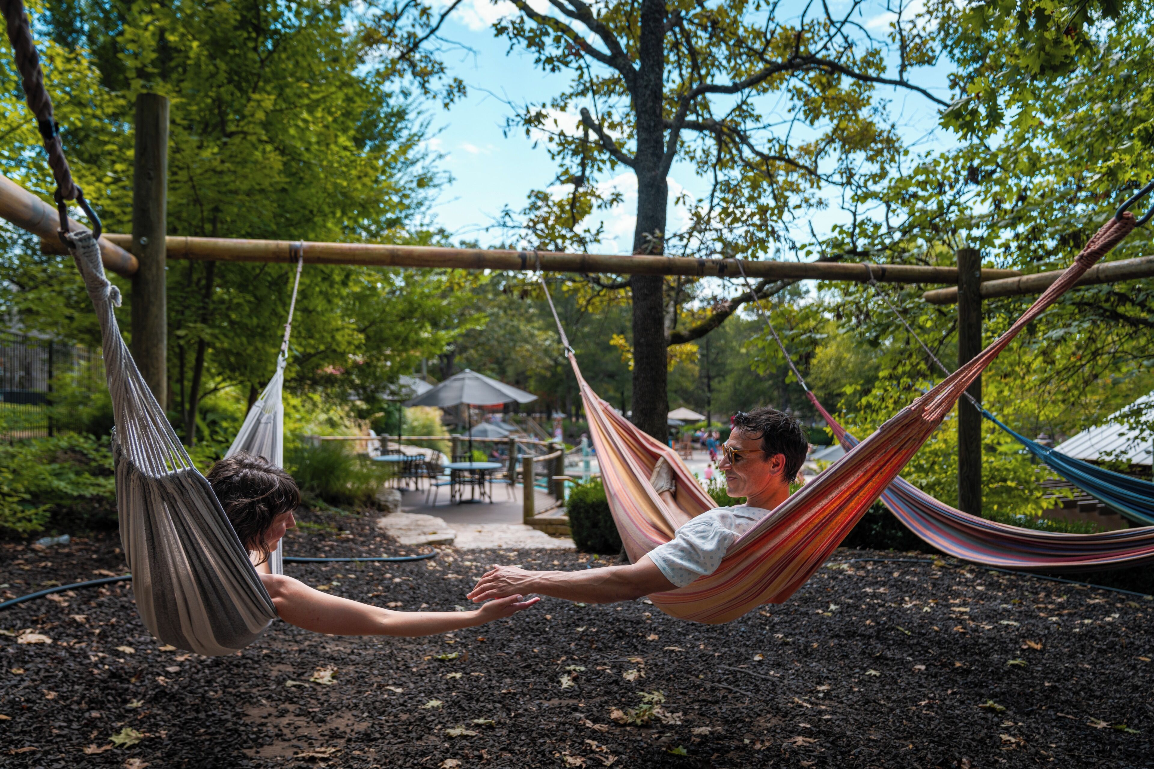 A hammock suspended between two poles in an outdoor setting with a table and chairs in the background.