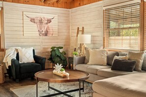 A cozy living room with a wooden wall, a large window with blinds, a black armchair, a grey sofa, and a coffee table with decorative items. - Spacious Resort Cabin – Sleeps 16, Minutes to Silver Dollar City! (Branson)