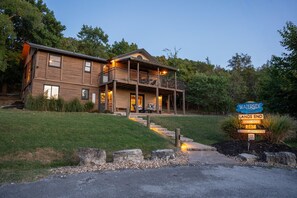 A two-story wooden cabin with a balcony, surrounded by a landscaped garden and a sign indicating a nightly rental. - Spacious Resort Cabin – Sleeps 16, Minutes to Silver Dollar City! (Branson)