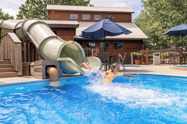 A water slide structure over a swimming pool with a wooden building in the background.