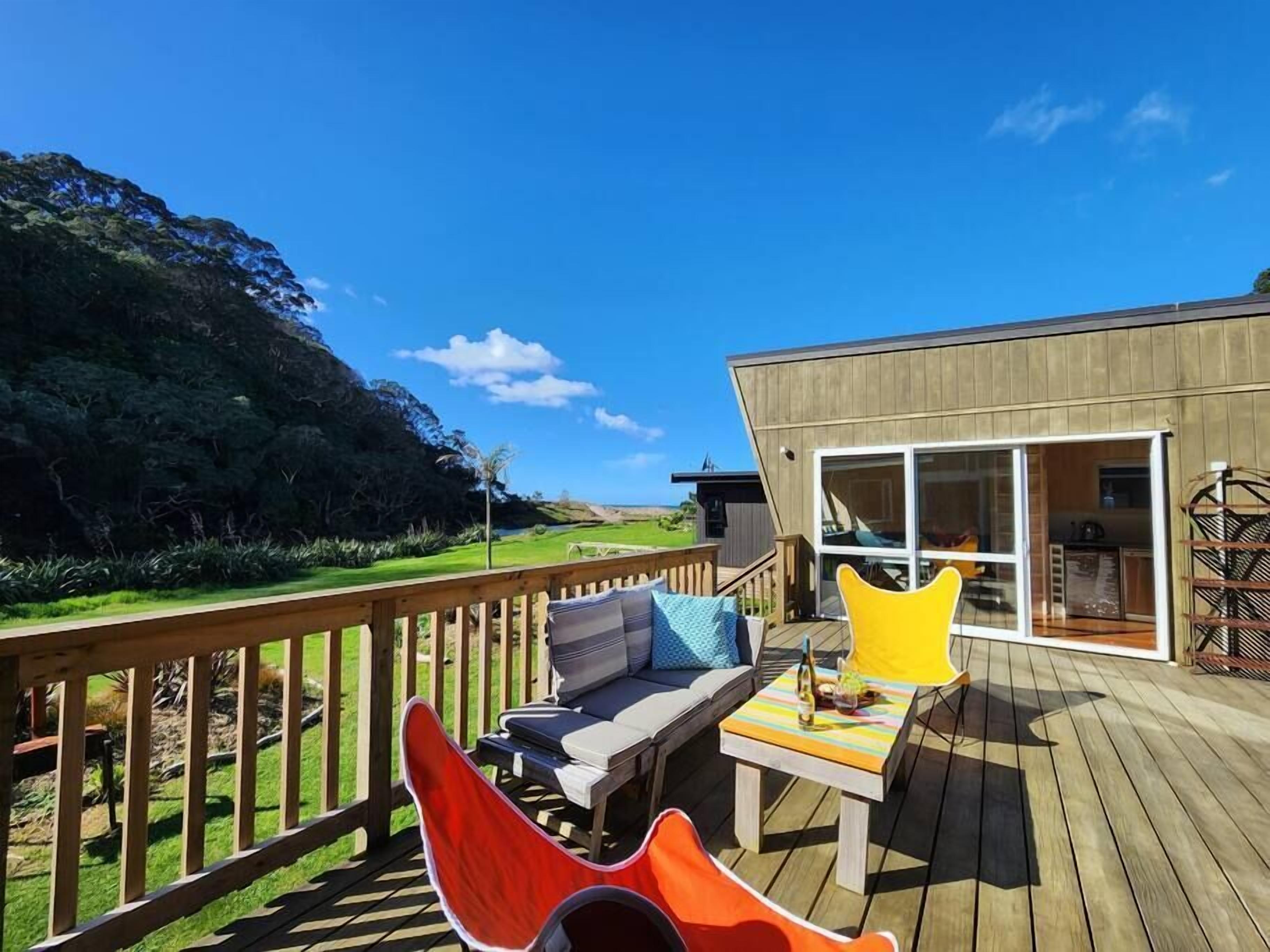 A wooden deck with a sofa and chairs, a table with a bottle, and a view of a mountainous landscape.