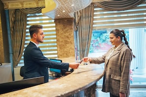 Balcão de recepção de hotel com um homem e uma mulher trocando um cartão, um monitor de computador e um telefone.