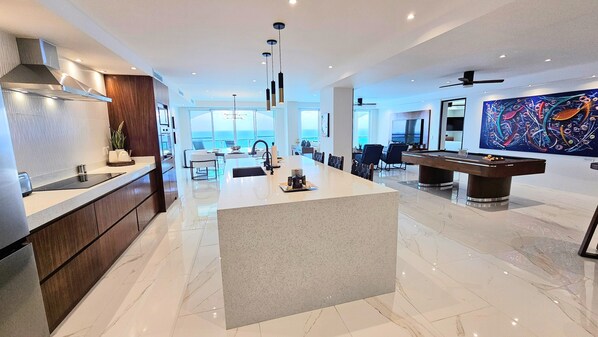 A modern kitchen with a central island, a dining area with chairs, a pool table, and a large window offering a view of the ocean.