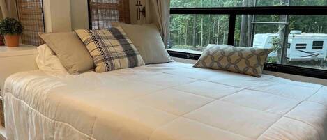 A large bed with a quilted cover, two windows with blinds, a potted plant, and a view of trees and a trailer outside.