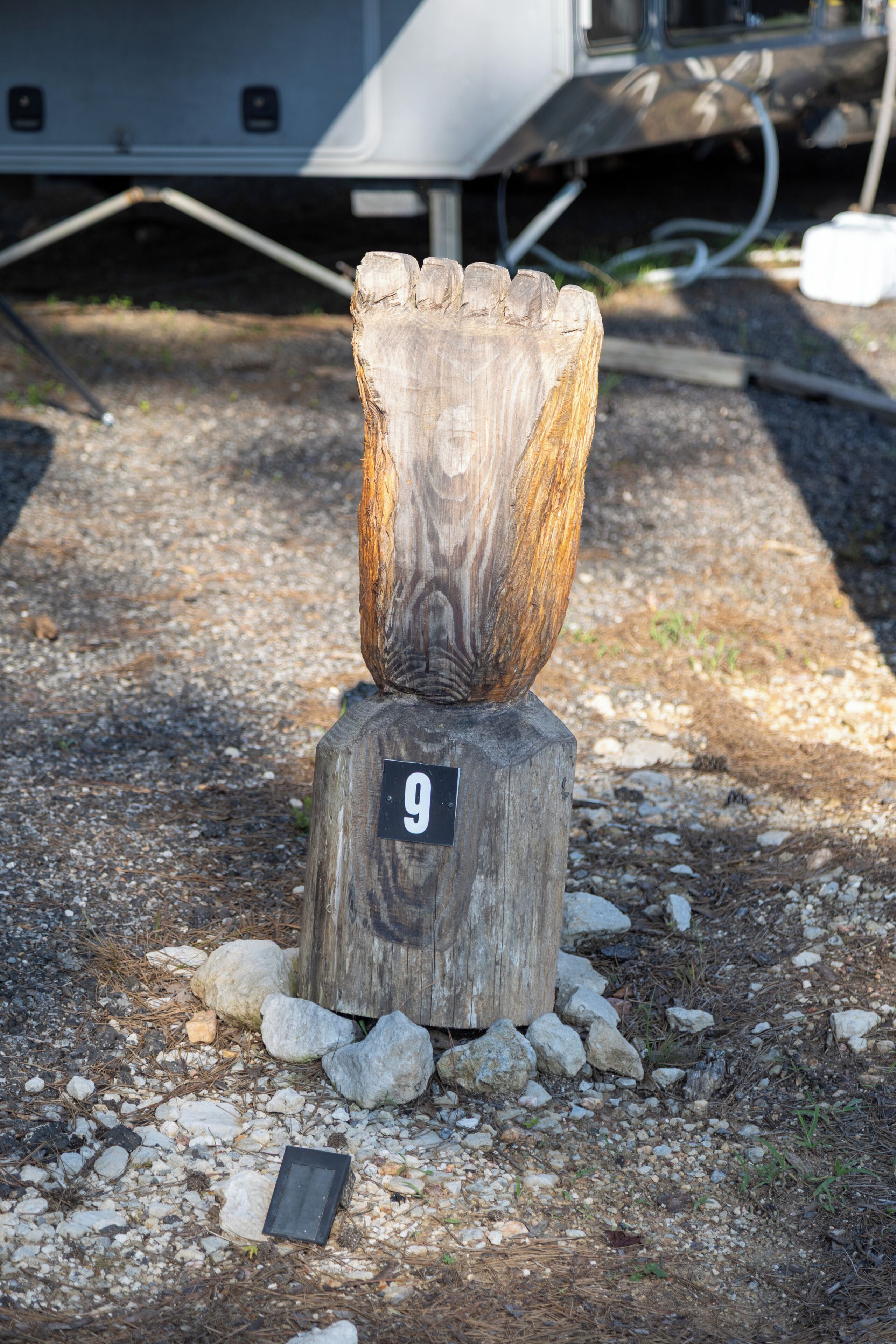 Une sculpture en bois représentant un visage sculpté, fixée sur un poteau en bois et portant le numéro 9. Elle est placée sur une surface de gravier, parsemée de roches, et un petit objet noir se trouve à proximité.