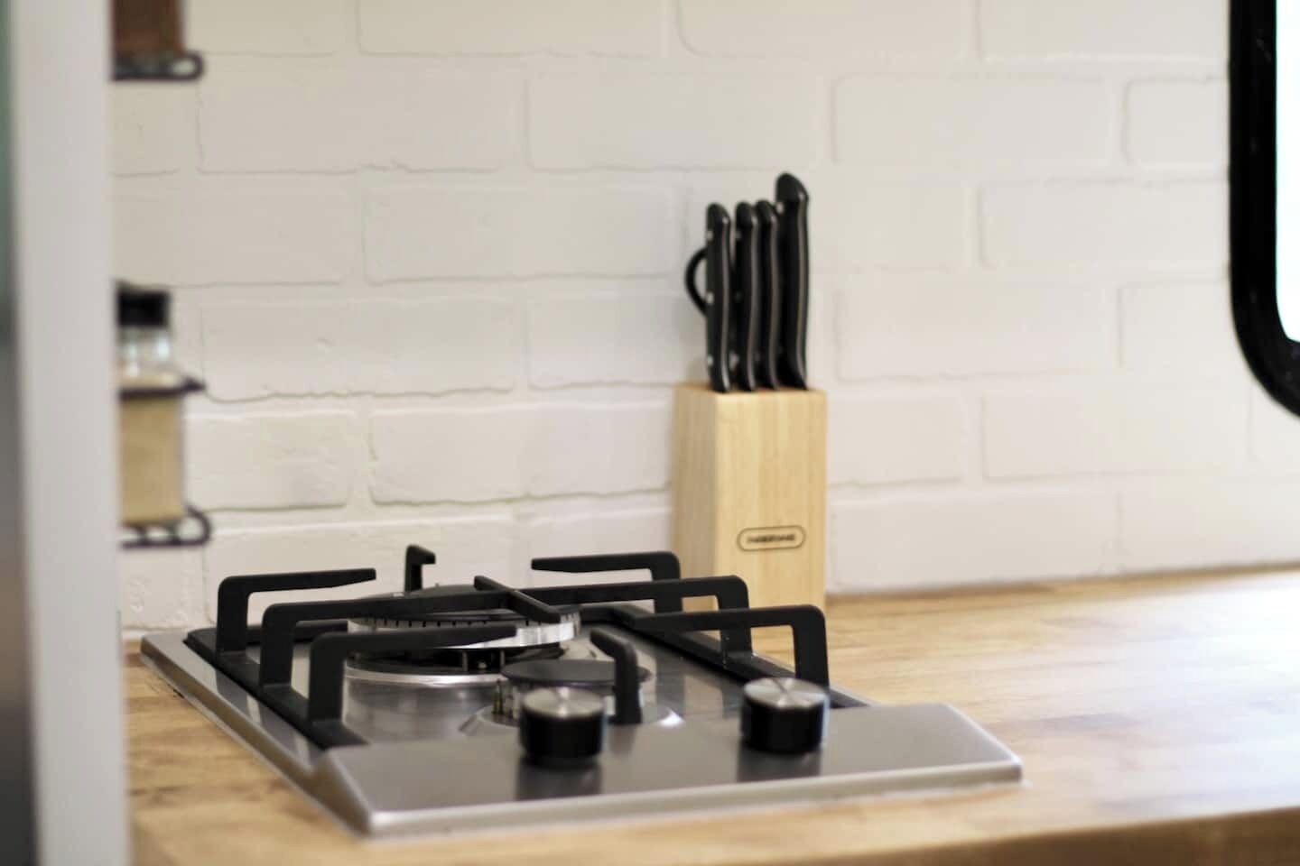 A kitchen with a gas stove, a knife block, and a white brick wall.