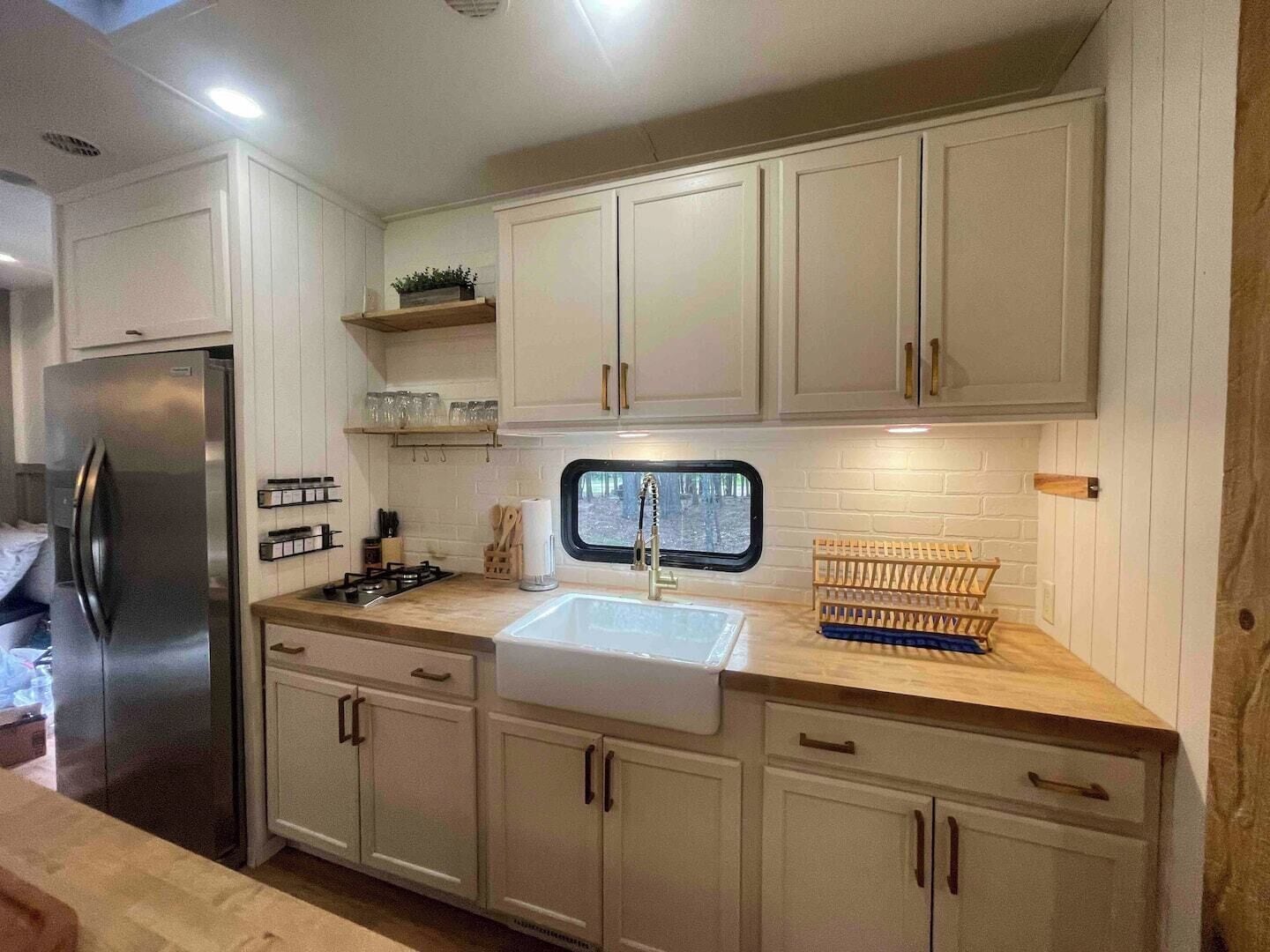A modern kitchen with white cabinets, a stainless steel refrigerator, a window above the sink, and a wooden countertop.