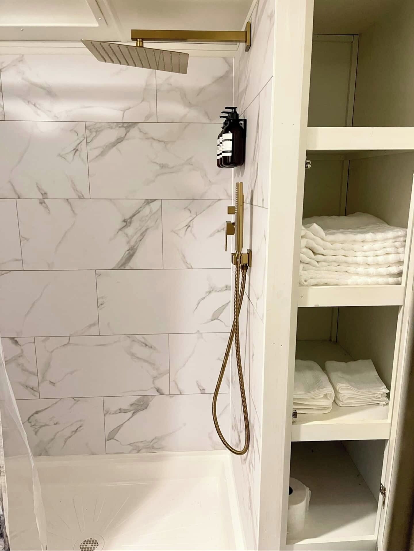 A bathroom with marble tiles, a gold showerhead, and a shelf with folded towels.