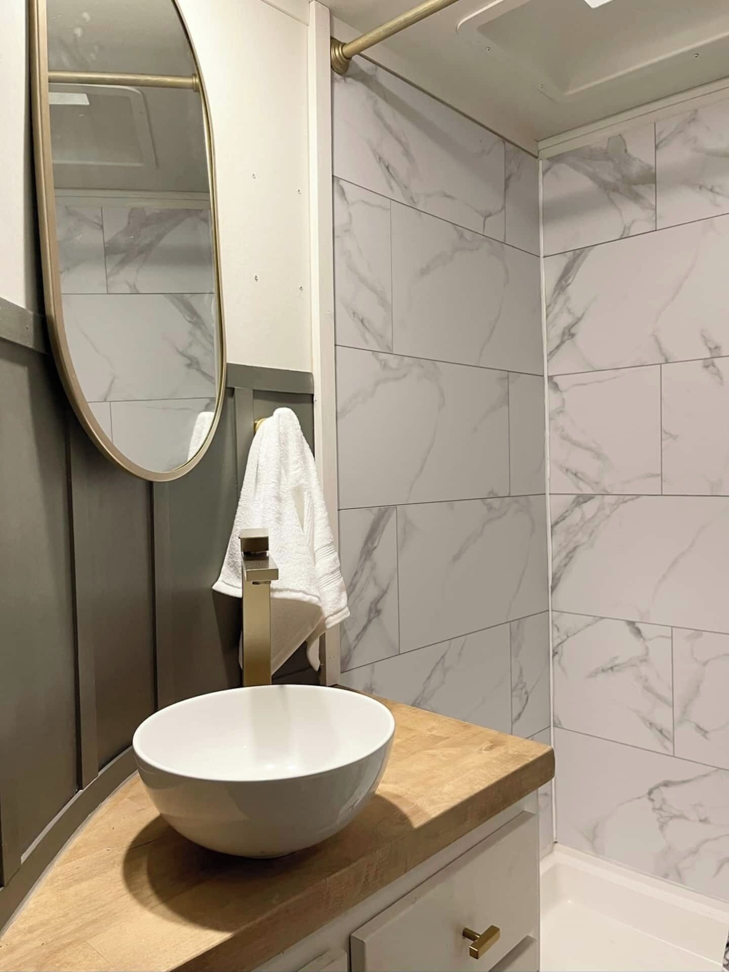 Un bagno con parete rivestita in marmo, lavandino bianco con rubinetto dorato e mobile contenitore in legno.