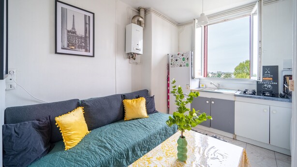 A modern living room with a sofa, a green quilted cover, and a yellow pillow. There is a framed picture on the wall, a window with a view, and a kitchen area with cabinets and a countertop. - L'Évasion Aulnaysienne - Appt pour 4 (Aulnay-sous-Bois)
