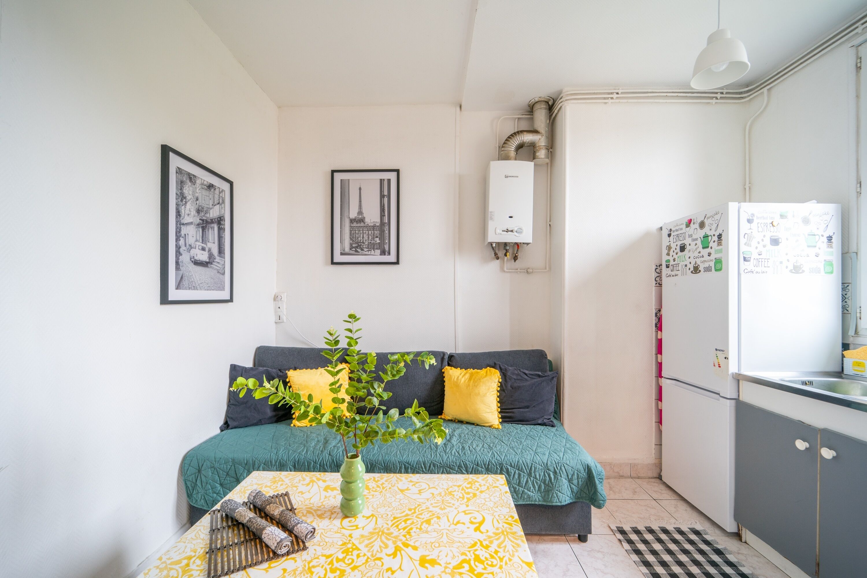 A small, modern apartment with a sofa, a coffee table with a vase, a refrigerator, and a wall-mounted heater.