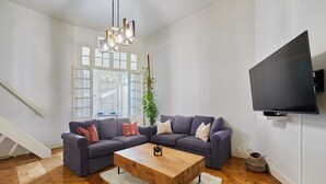 Un salon moderne avec une table basse en bois, des canapés gris, un tapis à poils longs blanc, une grande fenêtre avec des volets blancs et un téléviseur à écran plat fixé au mur.