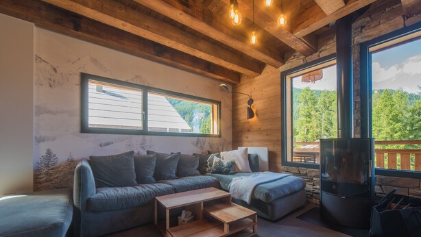 A cozy living room with a wooden ceiling, a large window with a mountain view, a fireplace, and a sectional sofa.