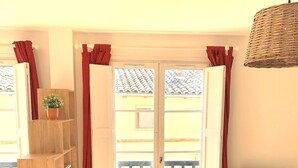 A bright living room with wooden flooring, a grey armchair with a red pillow, a TV stand, and a large window with red curtains. - Le Jaurès - Studio Apartment, Sleeps 2 (Toulouse)