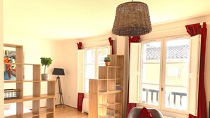 A room with a wooden floor, a chair with a red cushion, a floor lamp, a bookshelf, and a window with red curtains. - Le Jaurès - Studio Apartment, Sleeps 2 (Toulouse)