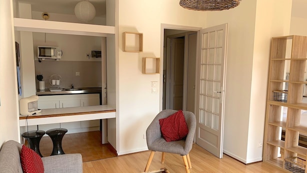 A modern living room with a sofa, a chair, a glass coffee table, and a kitchen area visible through an open door. - Le Jaurès - Studio Apartment, Sleeps 2 (Toulouse)