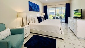 A modern bedroom with a large bed, a flat-screen TV, a ceiling fan, and a view of a balcony with outdoor furniture. - Regal Beach Club #311 (Seven Mile Beach)