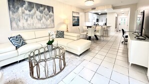 A modern living room with a white sofa, a glass coffee table, and a large abstract painting. - Regal Beach Club #311 (Seven Mile Beach)