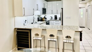 A modern kitchen with white cabinets, stainless steel appliances, and a granite countertop. There are three bar stools and a glass table. - Regal Beach Club #311 (Seven Mile Beach)