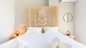 Ein ordentlich bezogenes Bett mit weißen Leinentüchern, zwei Nachttische mit Lampen, ein runder Spiegel und ein Traumfänger an der Kopfteil.