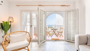 Ein helles Zimmer mit einem Korbstuhl, einer Couch und Blick auf eine Terrassenanlage mit Tisch und Stühlen.