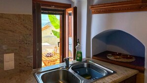 A kitchen with a double sink, a window with a view of an outdoor dining area, and a wooden shelf above the sink.