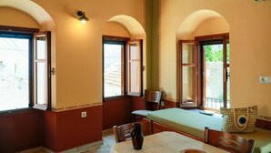 A room with a bed, a table, and a chair. There are windows with curtains, a vase on the table, and a wall with a decorative border.