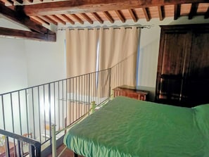 A bedroom with a green bed, wooden ceiling beams, a window with beige curtains, and a wooden door.