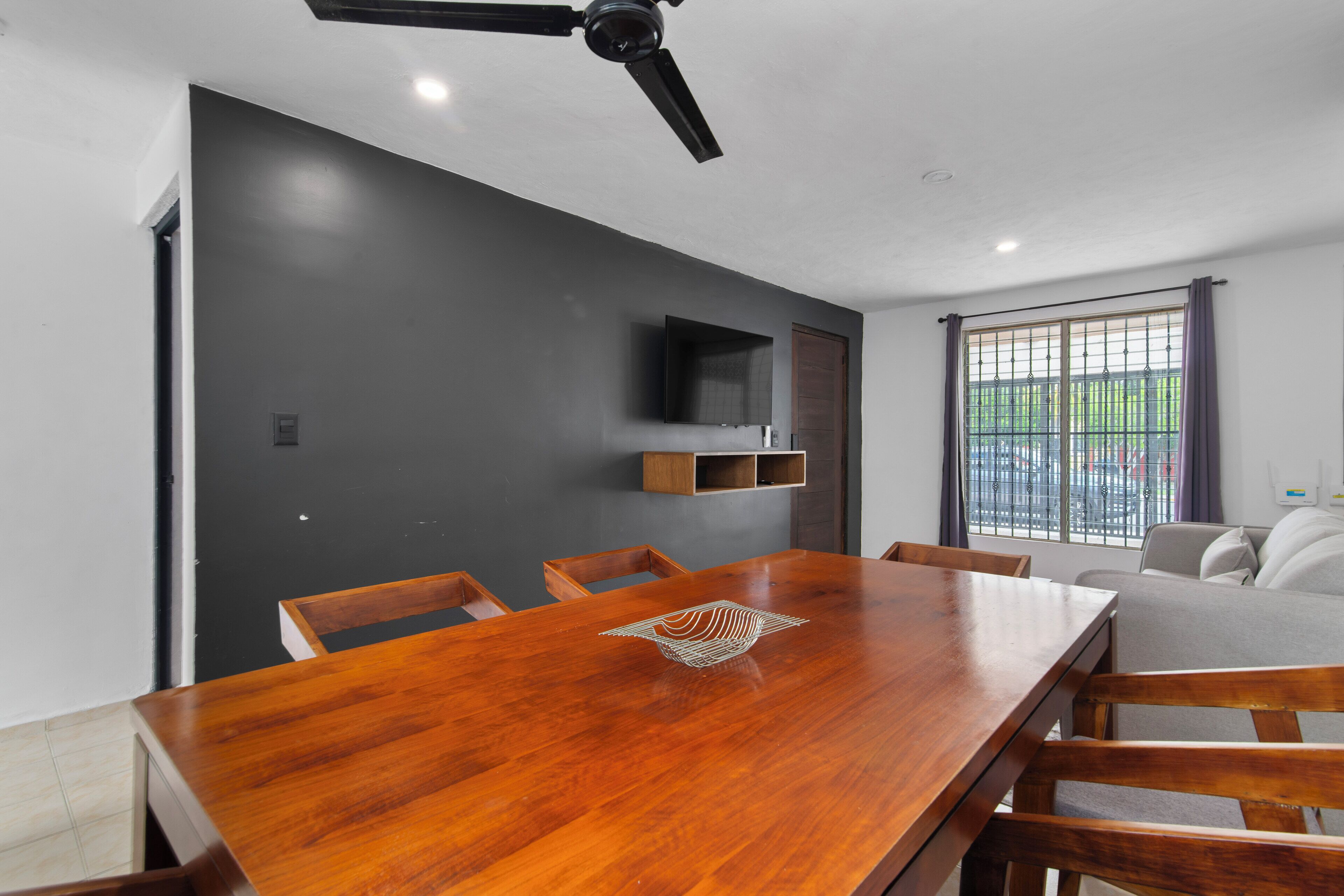 A modern dining area with a wooden table, chairs, a ceiling fan, a TV mounted on the wall, and a window with bars.
