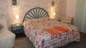 Een slaapkamer met een metselwerkmuur, een groen metalen hoofdbord, twee nachtkastjes met lampen, een bed met een geborduurd sprei en oranje handdoeken, een stoel en een kledingkast.