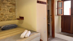 Una habitación con pared de piedra, techo de madera, una cama con toallas enrolladas, un estante pequeño con una copa de vino y una puerta que da a un baño.