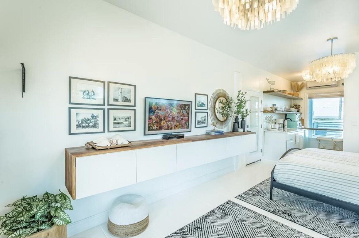 A modern bedroom with a bed, a console table with framed pictures, a chandelier, and a view of the kitchen.