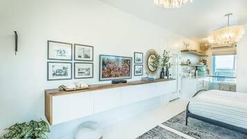 A modern bedroom with a bed, a console table with framed pictures, a chandelier, and a view of the kitchen.