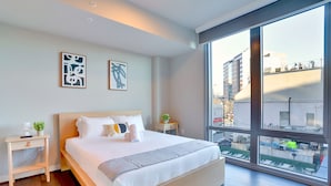 A modern bedroom with a large bed, two bedside tables, a nightstand, and a view of the city through large windows. - Cozy Apartments Near the Metro (Arlington)