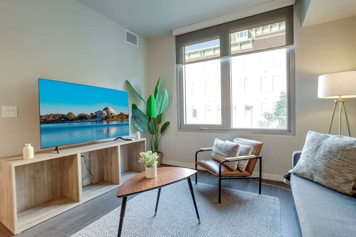 A modern living room with a large flat-screen TV, a wooden console table, a coffee table, a sofa, a chair, and a potted plant.