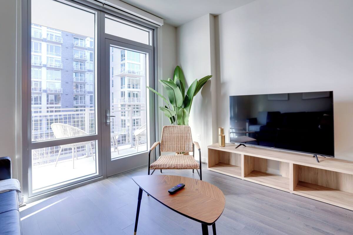 A modern living room with a large window, a flat-screen TV, a wicker chair, a wooden coffee table, and a potted plant.