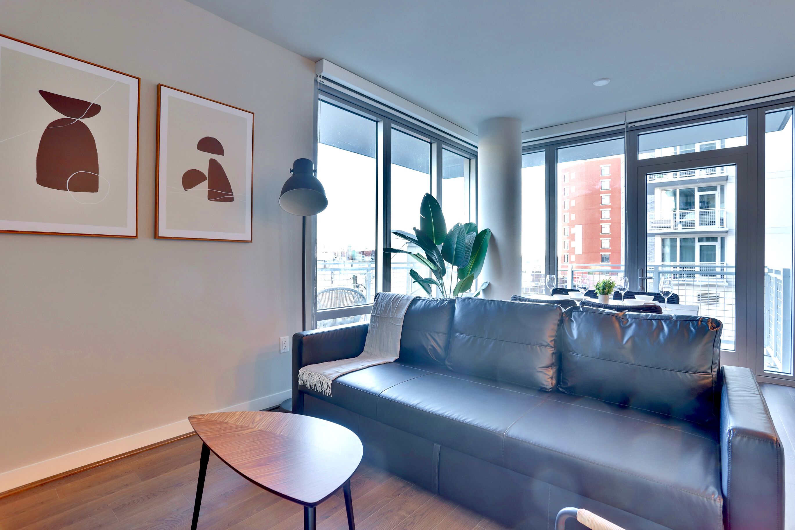 A modern living room with a leather sofa, a small wooden table, and large windows offering a city view.