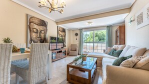 Una sala de estar con sofá, mesa de centro y sillas de mimbre. Se observan estatuas de Buda en la pared, un televisor y un reloj.