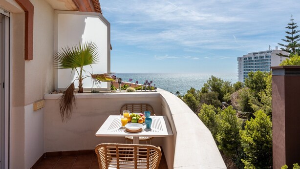 Un balcón con una mesa para dos, con vista al mar, una palmera y varias plantas cercanas.