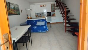 A compact kitchen with white cabinets, a dining table, and a blue patterned sofa. A wooden staircase is visible on the right. - Duna house in the green, relaxing terratet halfway between the sea and the countryside (Donoratico)