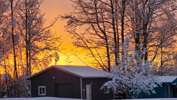 A snow-covered house with a garage, surrounded by trees and a sunset in the background. - At Paradise in the Valley (Palmer)